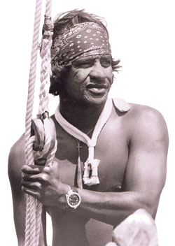 Eddie Aikau, shirtless, with a bandana around his head, holding onto the rigging for a boat. By http://www.honolulumagazine.com/archives/HON100/100Pictures/Eddie-on-Hokulea-c_o.jpg, Fair use, https://en.wikipedia.org/w/index.php?curid=12973191